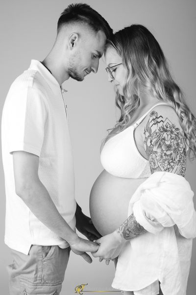 Ein schwangeres Paar, das beim Stehen vor einem grauen Hintergrund die Hände hält